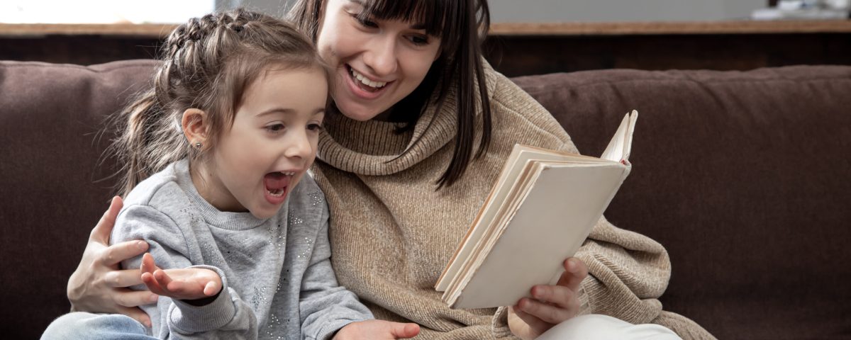 Young mother with her daughter are reading a book at home while sitting on the couch. Kind voorlezen 4 -6 jaar